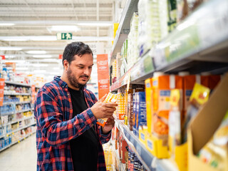 A man in a plaid shirt studies a product while standing in a grocery store aisle surrounded by...