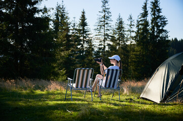 Woman camper relaxes next to tent with photo camera. Female tourist surrounded by tall evergreen trees and bathed in soft evening light, capturing peacefulness of moment.