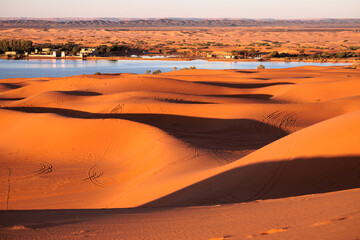 Sand dunes landscape at sunset, with traces of wheeled vehicles and Yasmina lake in the background. Erg Chebbi. Merzouga. Sahara desert. Morocco. Africa