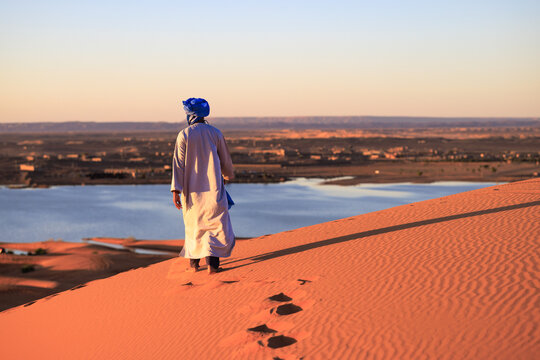 A tuareg in blue djellaba and turban, walking on the dunes of Erg Chebbi, with lake Yasmina in the background. Merzouga. Sahara desert. Morocco. Africa