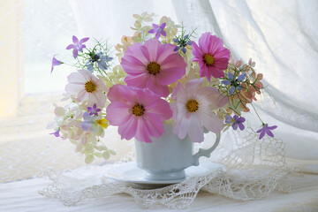 Bouquet of cosmos flowers, hydrangea, bells, pansies, nigella in a cup on a table by the window, a beautiful summer and autumn still life, postcard.