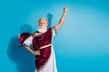 Photo of handsome senior male raise fist winning wear ancient greek god king dress isolated on blue...