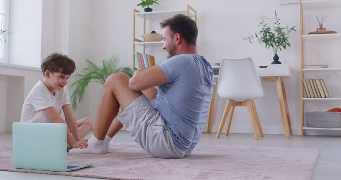 Little boy helping his father during routine fitness workout at home. Athletic man watching online video tutorial, training his abs by doing sit ups on floor with support of his son. Healthy family.