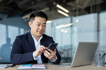 Smiling young Asian businessman sitting in office at desk with laptop and documents and using mobile phone