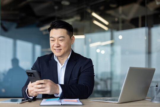 A young Asian man in a business suit is sitting in the office at the workplace and is using the phone, typing a message, making a call