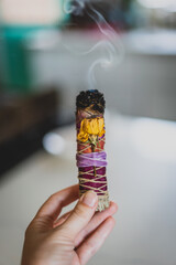 closeup of hand holding burning sage stick with smoke wrapped with flowers