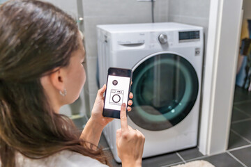 Young woman using mobile phone control via wi-fi and app for washing machine in home. Homesmart and new technology concept