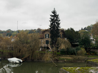 A serene riverside view featuring a historic house and a boat on a cloudy autumn day in a quaint town - Powered by Adobe