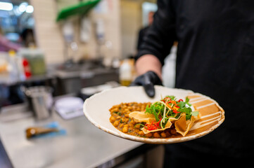 A close-up of a gloved hand holding a plate with a taco, garnished with fresh ingredients. The dish is presented in a casual dining setting, emphasizing the vibrant colors and textures of the food