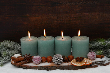 Vierter Advent: Arrangement mit grünen Kerzen, Tannenzweigen, Nüssen, Zimtstangen und Äpfeln vor einer Holzwand mit Platz für Text. 