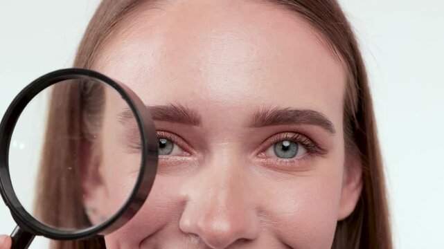Blonde woman with blue eyes on green background holding magnifying glass and showing her wrinkles on face