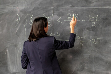 A female teacher of Asian descent writes algebra equations on a chalkboard, symbolizing education...