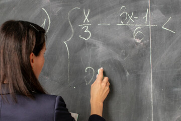 A female teacher of Caucasian descent solves a complex algebra equation on a chalkboard,...