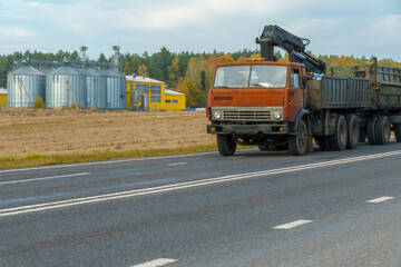 A truck is passing on the road in the background silver silos on agro manufacturing plant for processing drying cleaning and storage of agricultural products, flour. Large iron barrels of grain