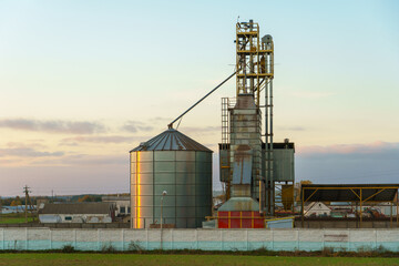 Obraz premium Grain silo elevator at sunset against the background of clouds. Iron barrels for grain storage. The harvest season.