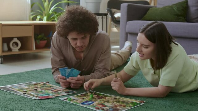 Young friends resting on green carpet in living room, smiling and enjoying strategy board game together at home