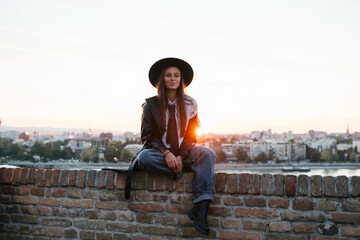 Fototapeta premium Young stylish woman enjoying sunset over city skyline from brick wall
