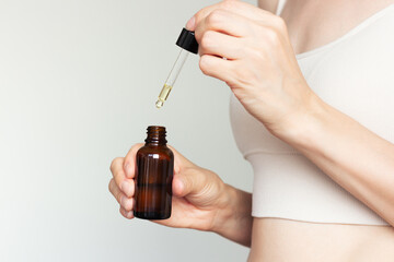 Profile shoot of young woman in beige underwear holding in her hand in front of her dark glass bottle of cosmetic liquid and dripping into it with pipette. Skin care. Isolated on a beige background