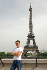 Stylish young male tourist stands on bridge against background of Eiffel Tower in Paris in capital of France. Photography, portrait, tourism concept.
