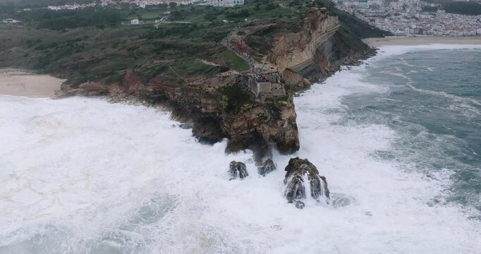 Aerial drone shot of big waves breaking on rocks and cliffs on a day with giant waves in Nazare, Portugal, Europe. Farol da Nazar&eacute; lighthouse visible. Shot in ProRes 422 HQ