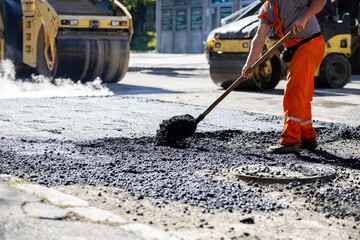 Construction worker laying asphalt on a city street during the day with road rollers in the background