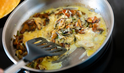 Process of cooking scrambled eggs in frying pan. Pieces of champignons and asparagus are added to dish. Hostess mixes dish with kitchen spatula, fry fried eggs with mushrooms