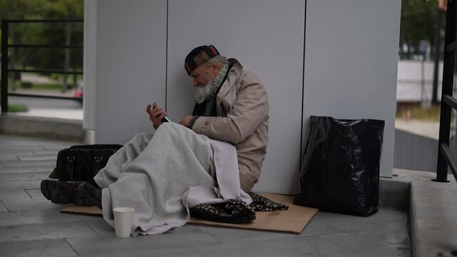 Desperate retired homeless man using smartphone sitting on cold winter street, wrapped in blanket for warmth, surrounded by meager belongings. Older tramp seek work and government assistance.