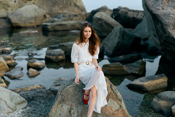 Serene woman in white dress sitting on ocean rock with feet in sand, feeling peaceful and connected to nature