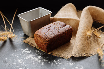 Rustic homemade rye bread loaf on burlap with wheat stalks and baking tin on dark background
