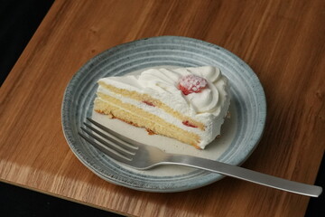 One small Japanese strawberry shortcake and a large fork on a stylish Japanese plate on a wooden tray on a black cloth on a white table