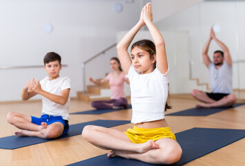 Cute tween girl sitting on mat in Padmasana yoga position with her hands clasped in prayer gesture above head while exercising yoga with brother and parents in fitness studio