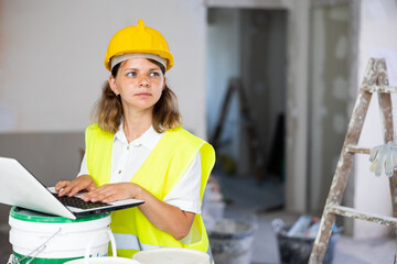 Female foreman in a protective helmet and a yellow vest checks the execution of repair work using a laptop