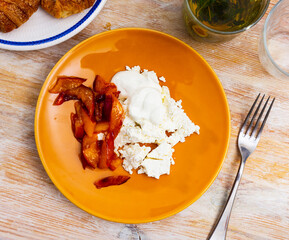 On white plate, portion of grainy cottage cheese with sour cream and peach slices in sugar syrup. Plate with croissants in background