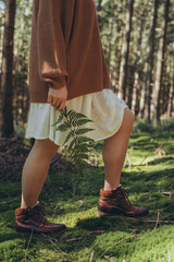 A woman surrounded by nature wearing a cozy and stylish outfit stands with a fern nearby