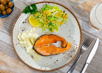 There is large piece of fried salmon on plate, garnished with mashed potatoes, vegetable salad, and portion of green olives.