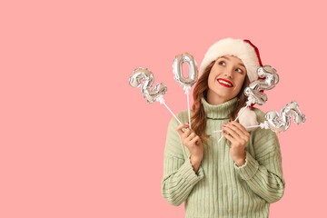Young woman in Santa hat with air balloons in shape of figure 2024 on pink background