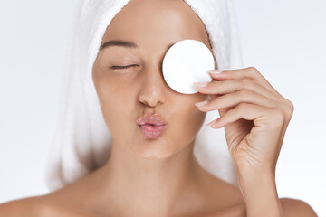 A playful portrait of a woman using a cotton pad to cleanse her face while puckering her lips. Wrapped in a towel, this image is perfect for skincare, beauty, and wellness promotions.