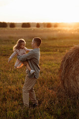 A man holds a young girl in a field. The sun is setting, casting a warm glow over the scene. Scene is peaceful and serene, as the father