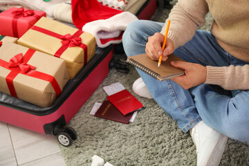 Young man with notebook sitting on floor near suitcase in living room. Christmas travel concept