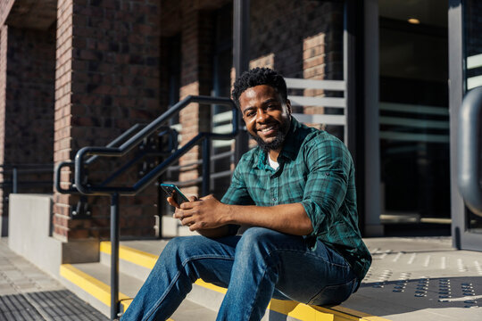 Smiling multicultural commuter is sitting at train platform on stairs with cellphone in hands.