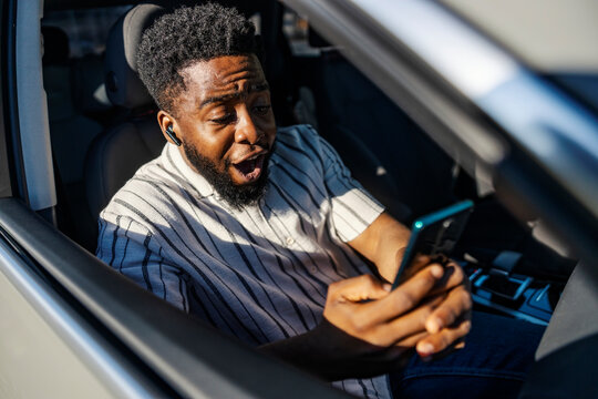Young happy black man sitting on passenger seat in car and getting excited about message on phone.