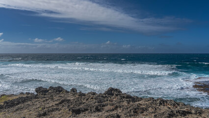 Endless blue ocean to the horizon. The waves are raging and foaming off the coast of the island. In the foreground is a rocky volcanic coast. Azure sky, clouds. Mauritius. Ile aux Fouquet's
