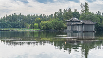 Fototapeta premium A Chinese-style building by a calm lake. The edges of the roof are curved. Reflection on the water. Lush green vegetation. Lotuses by the shore. Blue sky, clouds. China. Hangzhou West Lake Xi Hu 