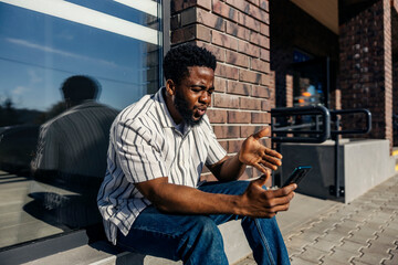 Nervous and angry black commuter sitting on bus platform with cellphone in hands and waiting for bus