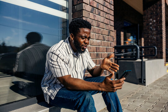 Nervous and angry black commuter sitting on bus platform with cellphone in hands and waiting for bus