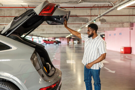Young black man is opening car trunk at garage and looking for spare tire.