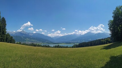 Obraz premium Panoramic view of a mountain lake with green meadow in the foreground and a clear blue sky with clouds.