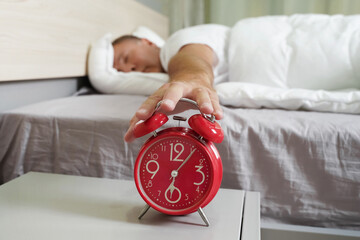 Person reaching for a red alarm clock while lying in bed in the morning light