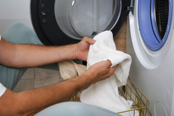 A man sits comfortably on the floor beside a washing machine, sorting socks from a basket in a well-lit laundry room during the afternoon.