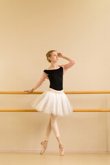 A focused ballerina stands gracefully on pointe, holding the barre with one hand and looking upward